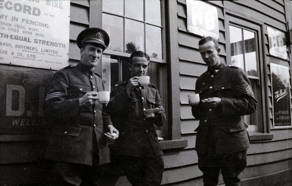 Norman Shepherd (right) and two other sergeants stand outside a wooden building at Kaitoke, drinking tea.